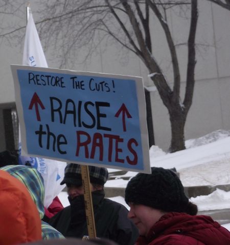 Demonstrators and a sign demanding an increase to the social assistance rates, during the portion of the Solidarity Against Austerity event that happend outside the provincial building in downtown Sudbury. (Photo by Scott Neigh)