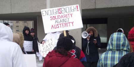 Demonstrators and a "Solidarity Against Austerity" sign outside the provincial building. The speaker is Menal Mehari of Laurentian University's Graduate Student Association and the Canadian Union of Public Employees Local 5011. (Photo by Scott Neigh)